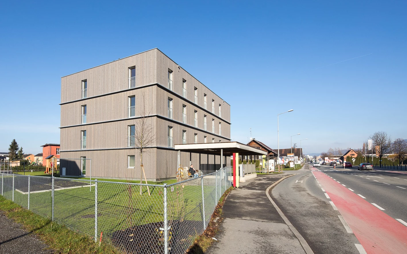 Modernes Mehrfamilienhaus mit Holzfassade und Balkonen am "Froschweg" in Dornbirn. Vor dem Gebäude befindet sich eine Parkgarage, ein Spielplatz und ein Gehsteig neben einer Straße.