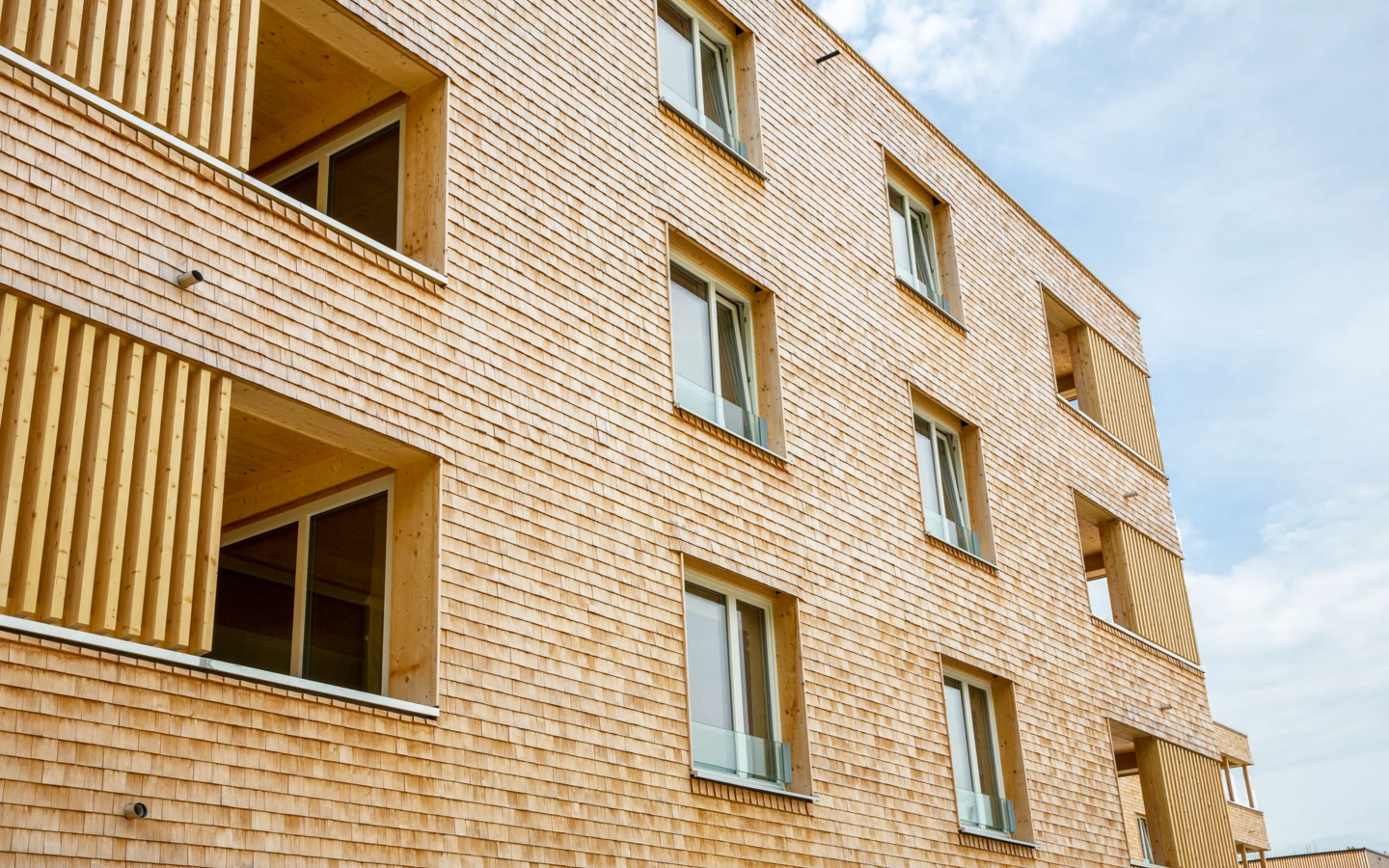 Fassadenansicht eines modernen Gebäudes mit Holzschindelverkleidung und großen Fenstern sowie hölzernen Lamellen an den Balkonen (Hellbrunnenstraße).