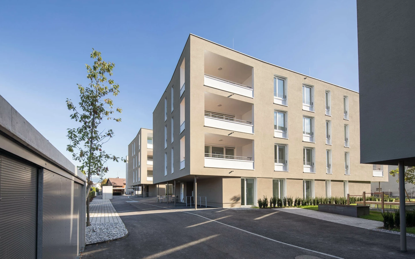 Wohnanlage Widum Lustenau 14 mit modernen Fassaden, Balkonen und bodentiefen Fenstern, umgeben von asphaltierten Wegen und minimalistischer Bepflanzung.
