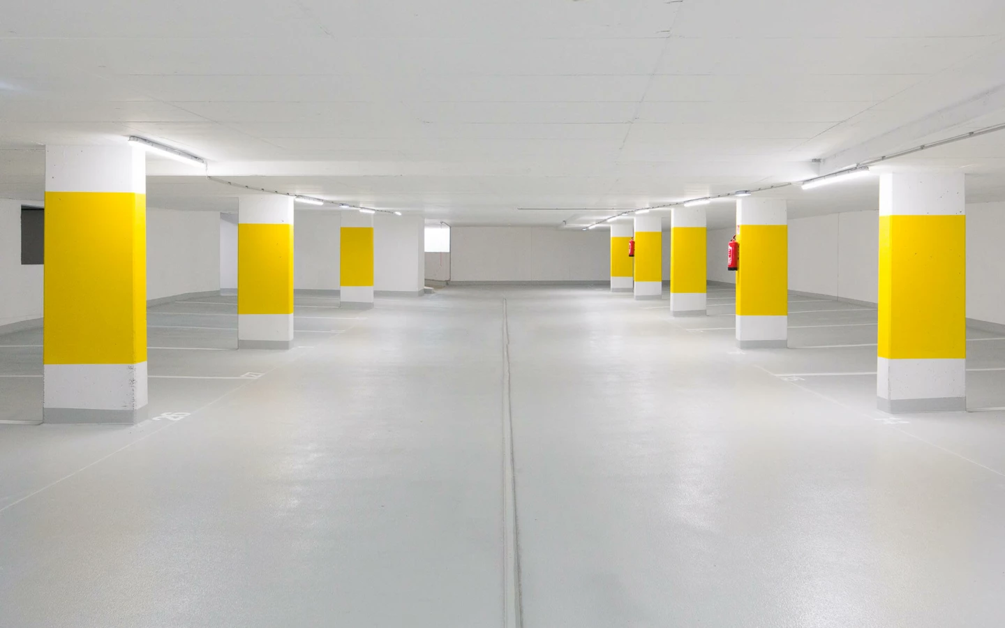 Leere Tiefgarage mit grauer Bodenfläche, weißen Wänden und gelb-weiß gestrichenen Säulenreihen mit Parkplatzmarkierungen (Widum, Wohnanlage, Lustenau).