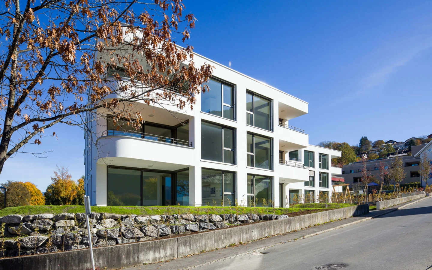 Moderne Wohnanlage Wolf-Huber-Straße in Feldkirch mit weißer Fassade, großen Fenstern und Balkonen an einem sonnigen Herbsttag.