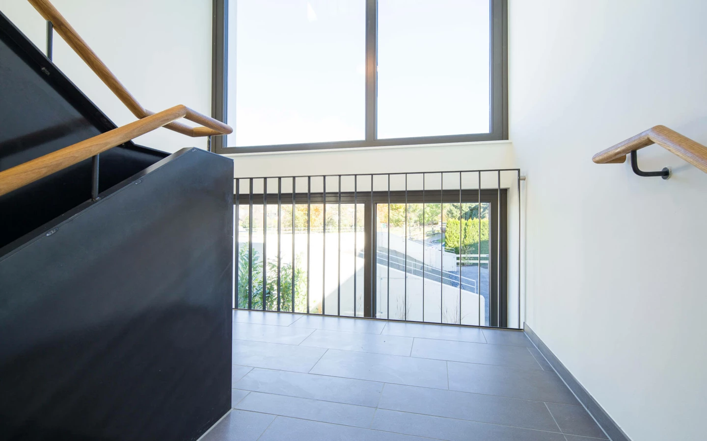 Moderner Treppenbereich mit dunkler Treppe, Holzhandläufen und großer Fensterfront in der Wohnanlage Wolf-Huber-Straße, Feldkirch.