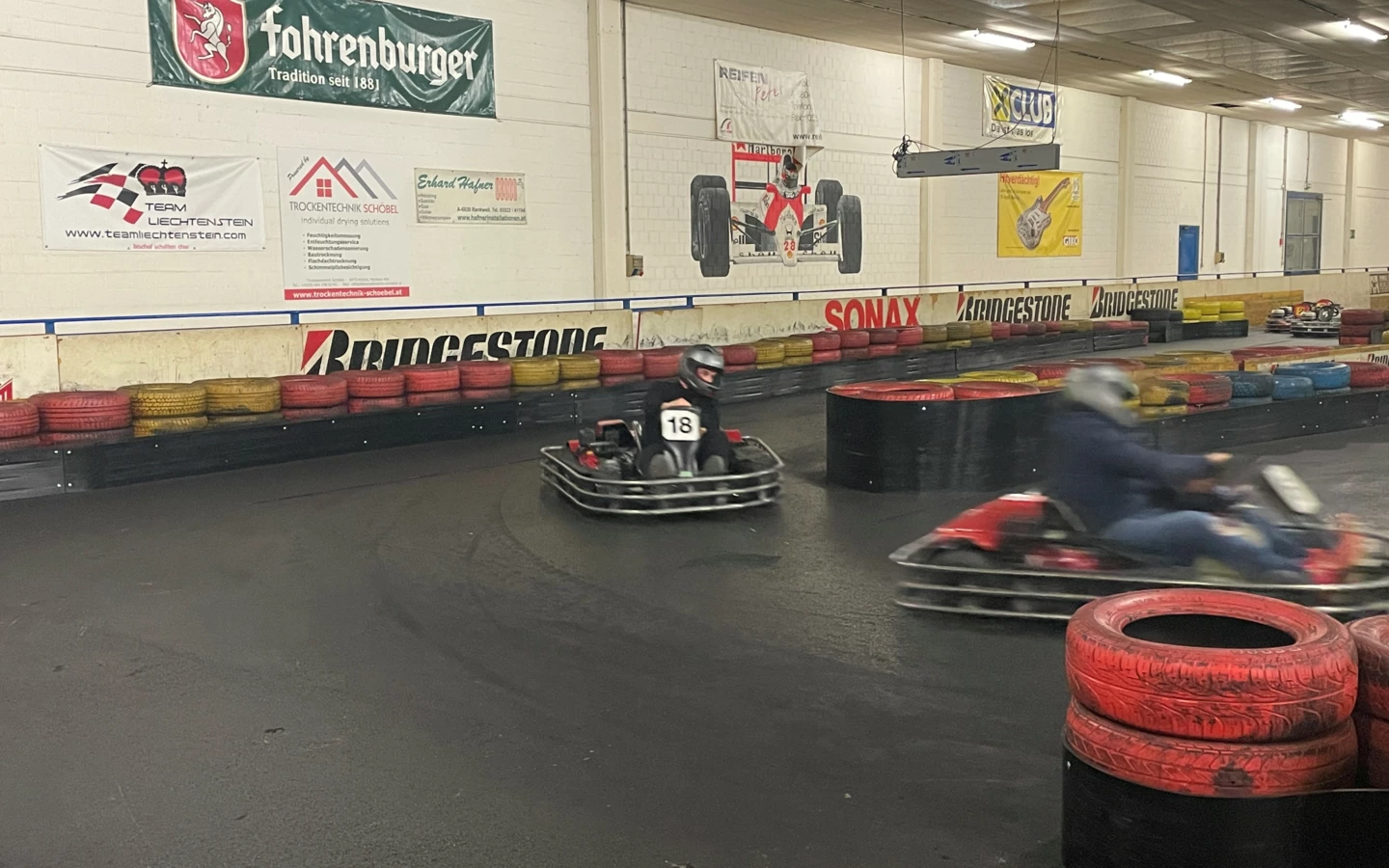 Zwei Personen fahren Go-Karts auf einer Rennstrecke mit Reifenstapeln und Werbebannern an den Wänden (Lehrlingsabend).