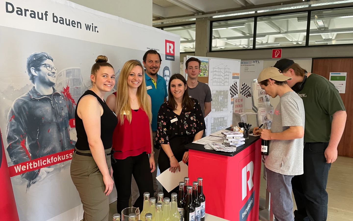 Mitarbeiter von Rhomberg Bau an einem Messestand, der "Darauf bauen wir" und "Weitblickfokus" bewirbt mit Getränken und Broschüren (Bauforum).