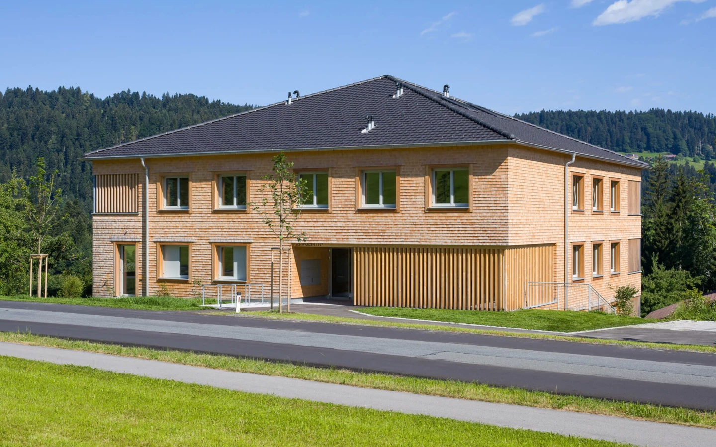 Modernes Wohnhaus mit Holzfassade und dunklem Dach, umgeben von grünen Wiesen und Wäldern unter blauem Himmel in Schwarzen, Alberschwende (Wohnanlage).