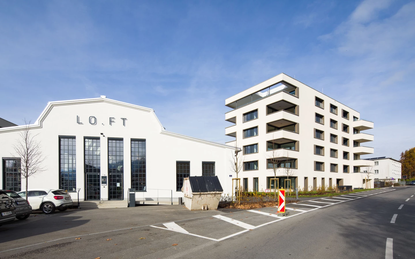 Zwei moderne Gebäude, eines mit "LO.FT" Schriftzug und großen Fenstern, sowie ein Wohnhaus mit Balkonen an einer Straße bei Tag (Bäumleareal).