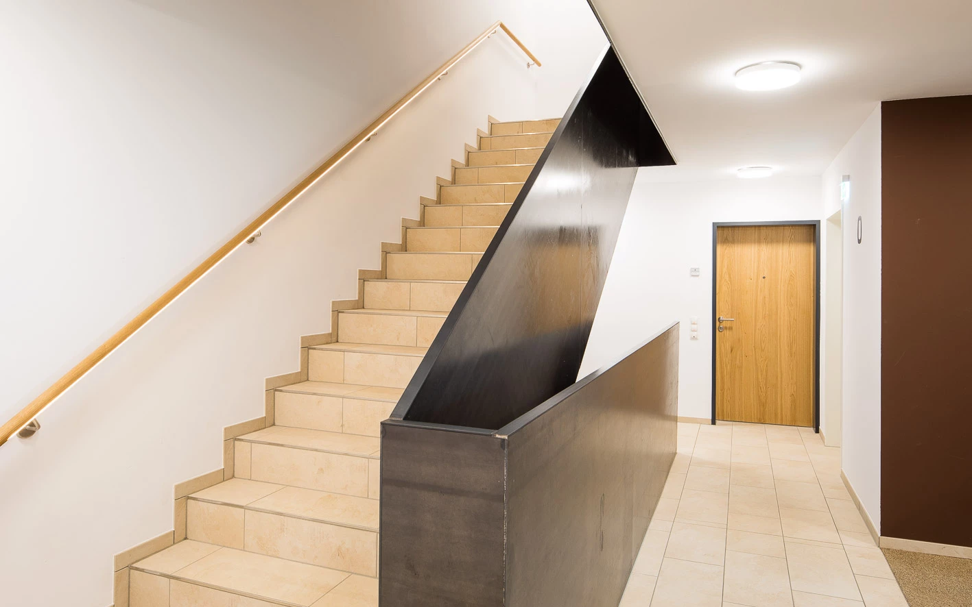 Ein modernes Treppenhaus mit hellen Fliesenstufen, einem Holzhandlauf und einem dunklen Metallgeländer. Eine Holztür ist am Ende des Flurs sichtbar (Baeumlequartier).