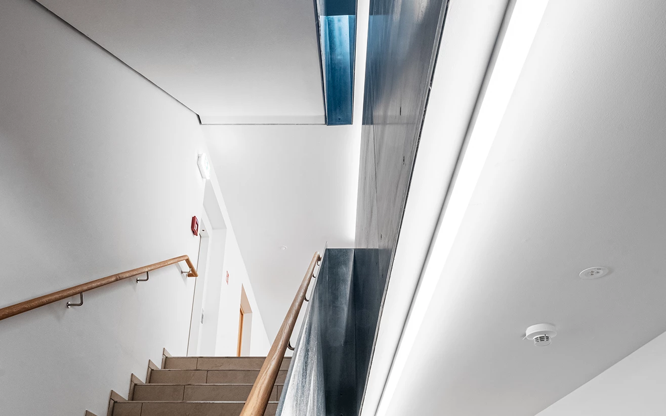 Eine breite helle Treppe mit beigem Steinbelag und Holzgeländer führt nach oben, beleuchtet von langen LED-Streifen (Seeblick, Lochau).