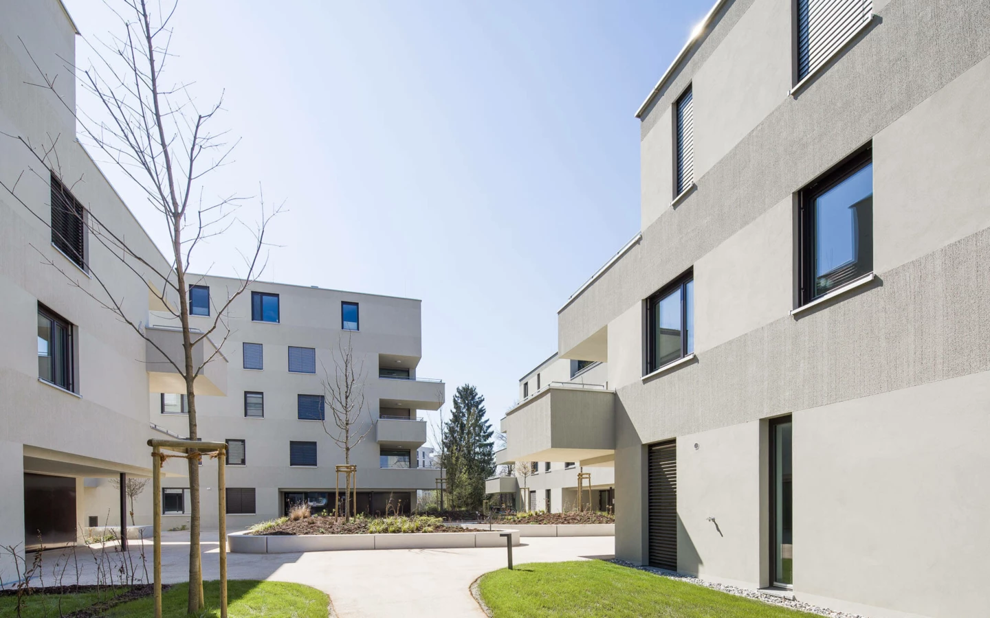 Moderne Wohnanlage Wuhrbaumweg von Rhomberg Bau mit hellen Fassaden, Balkonen und gepflegten Grünflächen unter blauem Himmel.