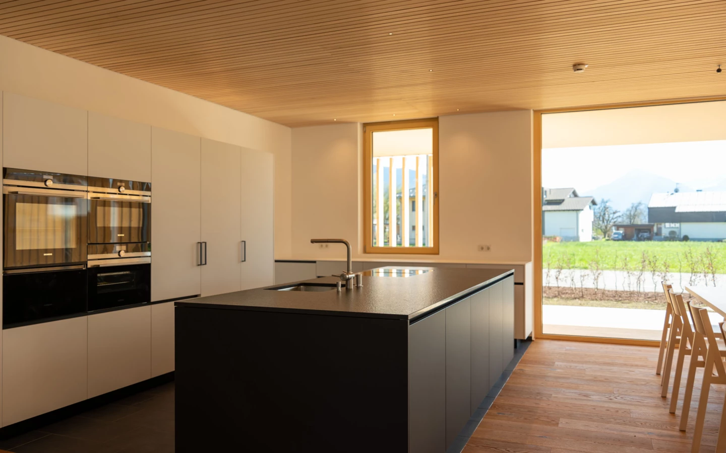 Moderne Küche mit schwarzer Kücheninsel, weißen Schränken und brauner Holzdecke. Große Fensterfronten bieten Blick auf grüne Wiesen und Berge (Wolfurt, Dieter).