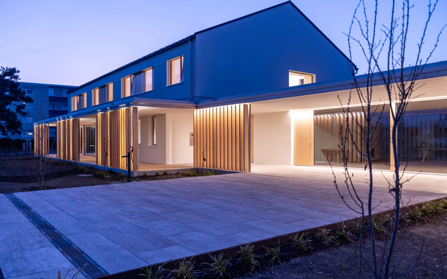 Modernes Wohnhaus mit blauer Fassade und beleuchteter Holzverkleidung im Eingangsbereich, abends aufgenommen (Wolfurt, Dieter).