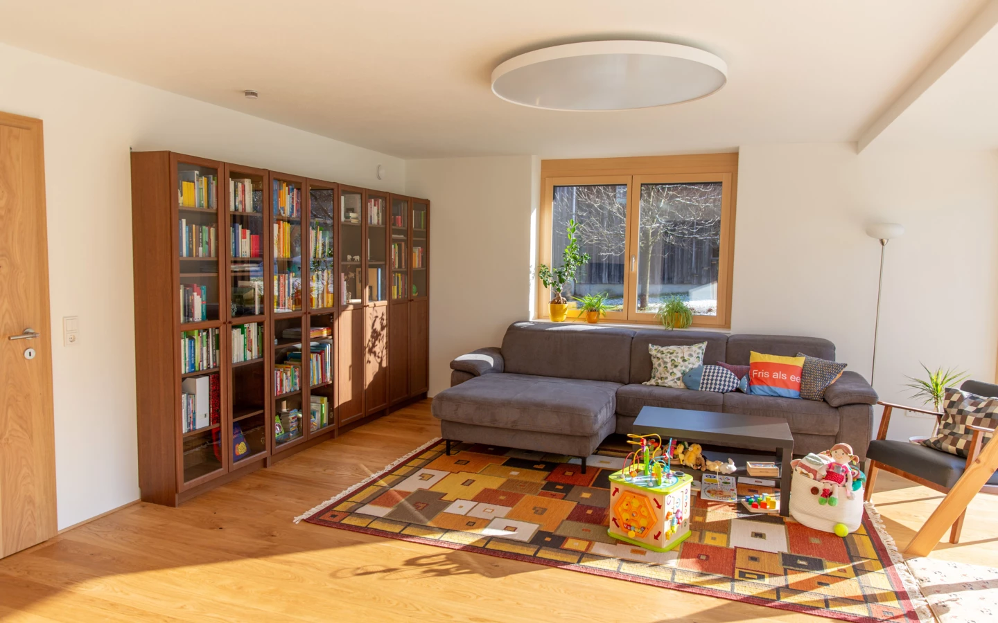 Helles Wohnzimmer mit L-förmigem Sofa, Bücherregal und Teppich. Sonnenlicht fällt durch das Fenster (Generalsanierung, Einfamilienhaus, Holzoptik, Dornbirn).