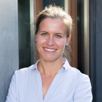 Eine lächelnde junge Frau mit hochgesteckten Haaren und einem hellblauen Hemd lehnt an einer grauen Säule (Manuela, Fetz).