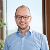 Christoph Sorg lächelt in die Kamera, trägt ein hellblaues Hemd und eine Brille, mit einer Pflanze und Fenster im Hintergrund.