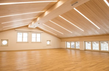 Ein großer, leerer Raum mit Holzboden und einer schrägen Holzdecke, die mit langen, linearen LED-Leuchten versehen ist (Generalsanierung, Kindes, Lech).