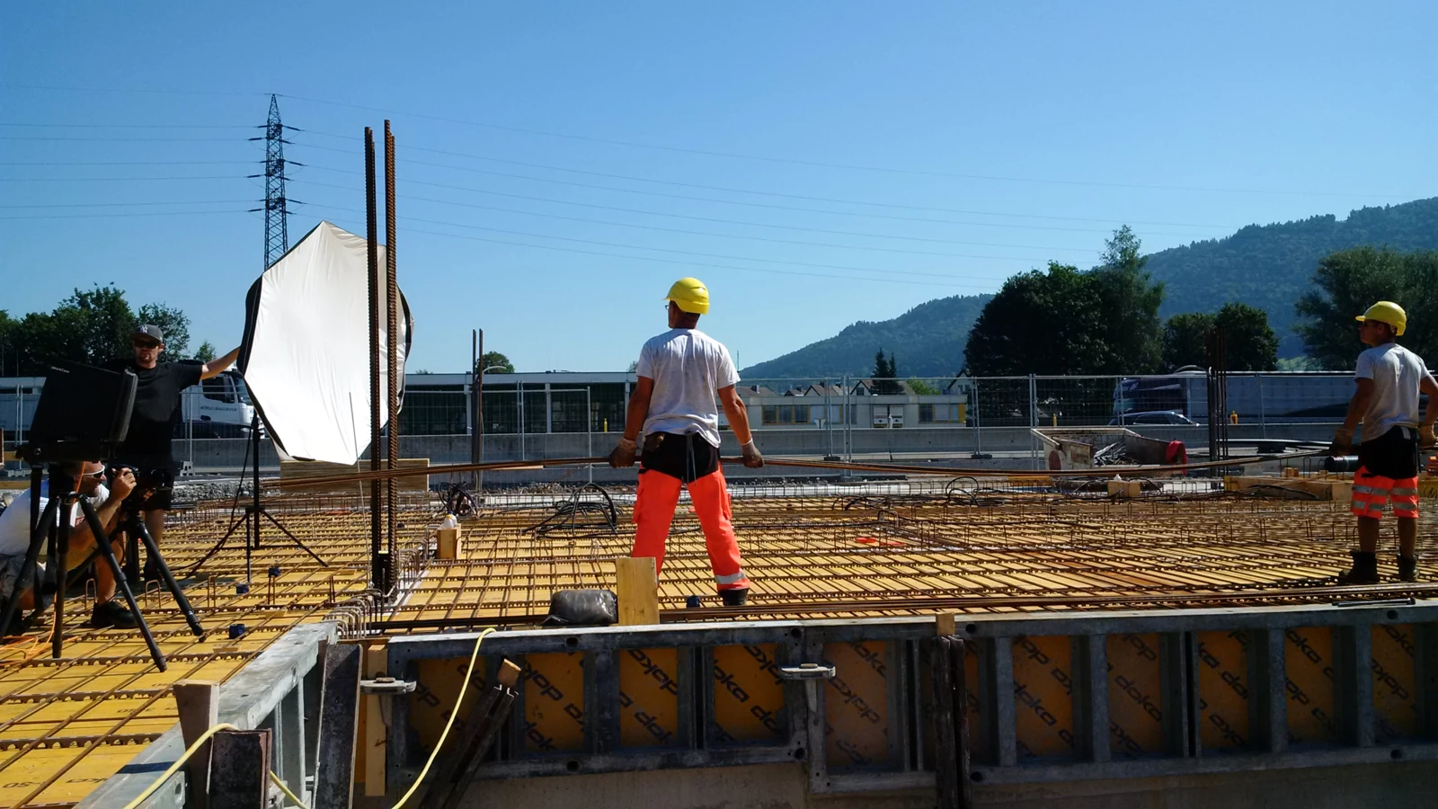 Zwei Bauarbeiter in Schutzkleidung und ein Fotograf mit Softbox auf einer Baustelle unter blauem Himmel mit Bergen und einem Hochspannungsmast im Hintergrund.