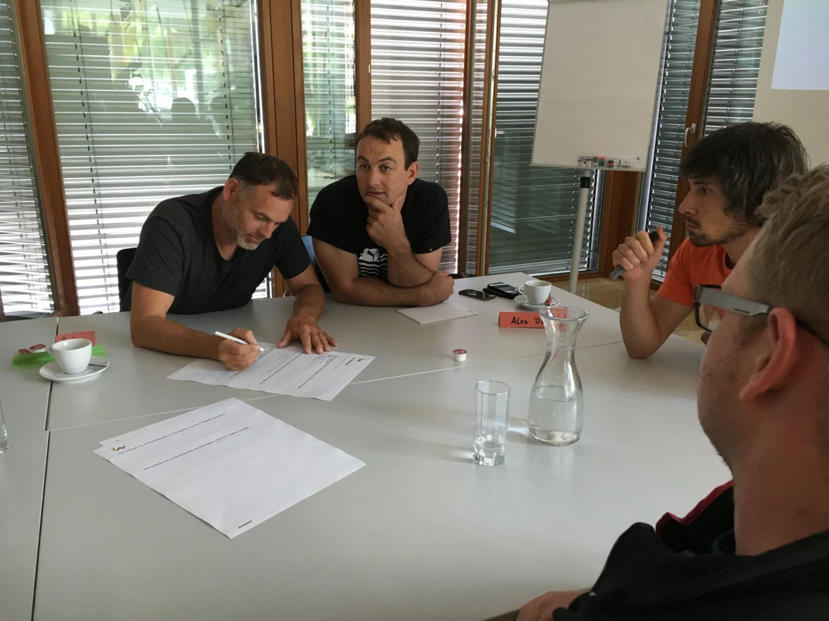 Vier Männer sitzen an einem Tisch und diskutieren bei einem Workshop, einer davon schreibt Notizen auf ein Blatt Papier.