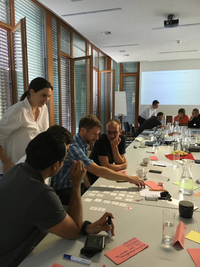 Eine Gruppe von Rhomberg Bau Mitarbeitern sitzt an einem Tisch bei einem Workshop und arrangiert Karten. Eine Präsentation ist im Hintergrund an die Wand projiziert.