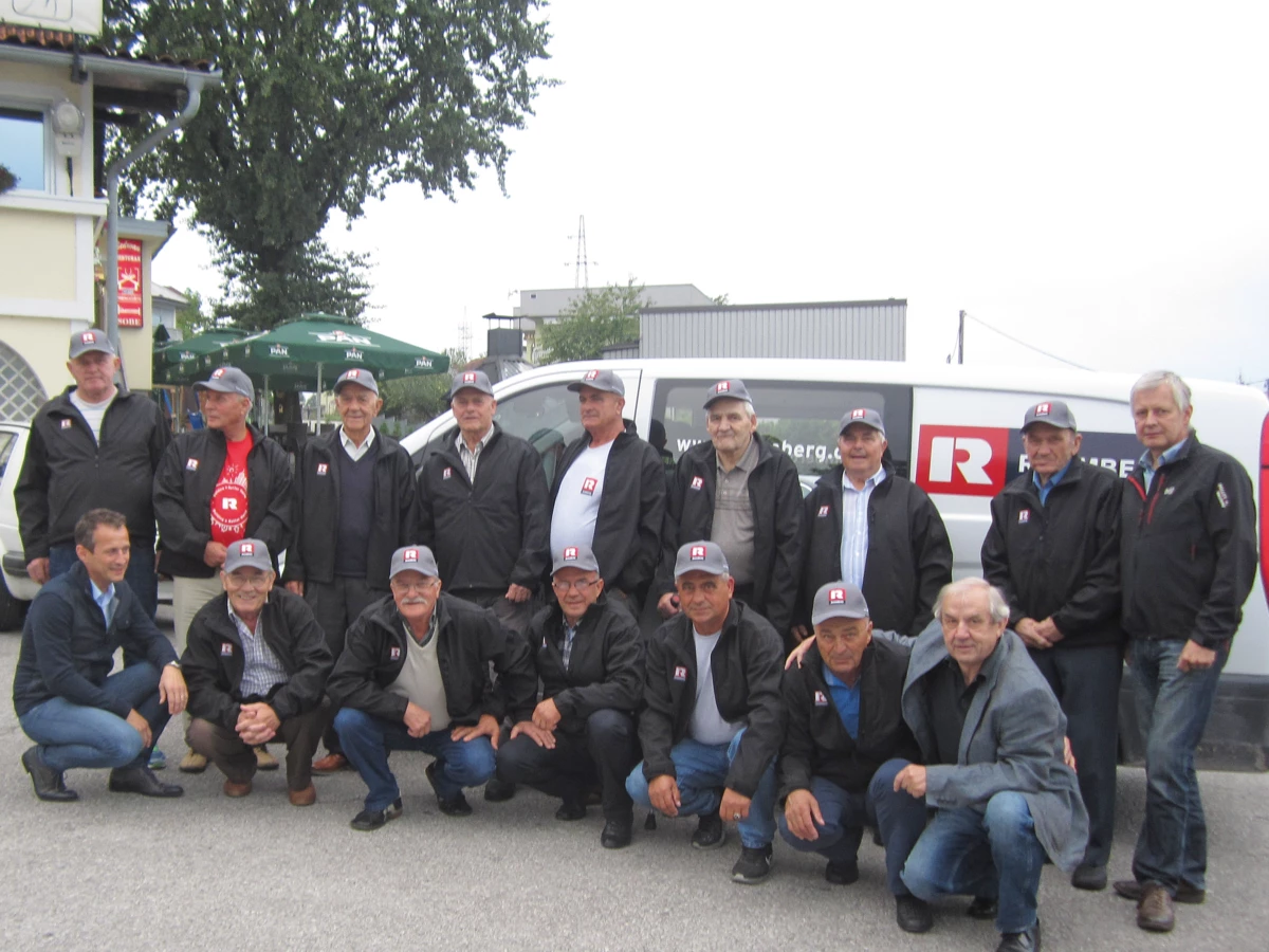 A group of eighteen men of various ages, most wearing black jackets and gray Rhomberg caps, pose outdoors. Some crouch in front of a white (pensioners)