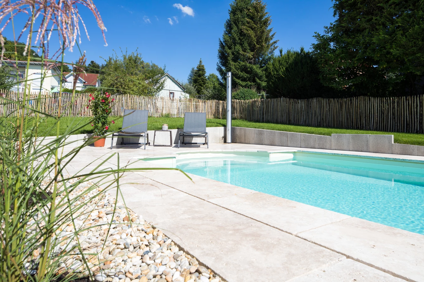 Ein rechteckiger Pool mit hellblauem Wasser, umgeben von beigem Steinbelag und zwei grauen Liegestühlen, an einem sonnigen Tag (Hohenweiler).