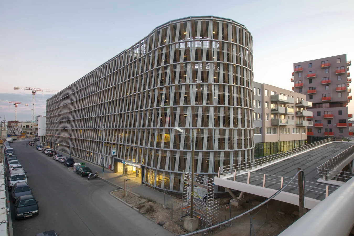 Rundes Parkhaus Wien Hauptbahnhof mit Holzlamellenfassade bei Dämmerung, umgeben von Wohngebäuden und einer Straße mit parkenden Autos (Hochgarage).