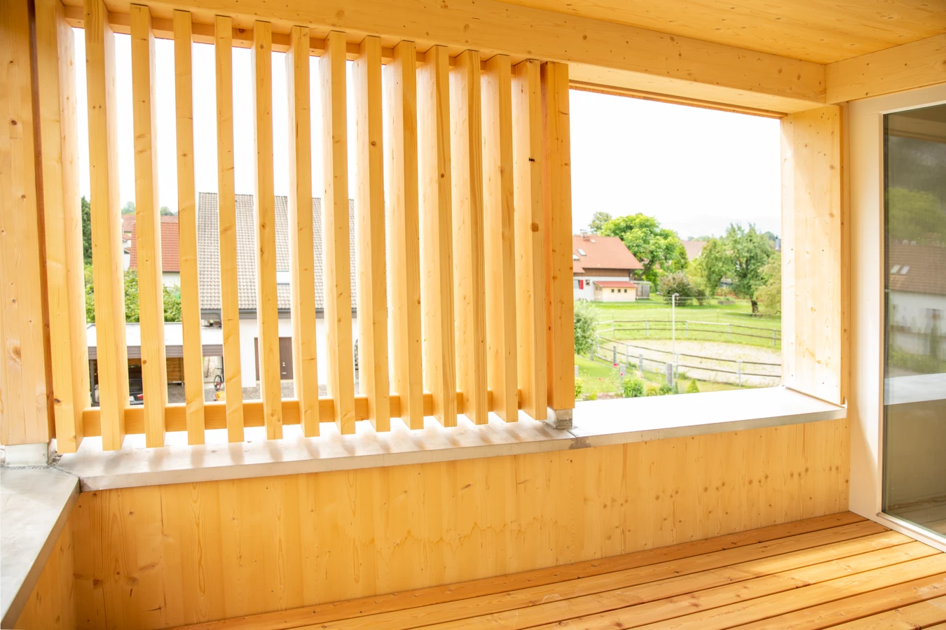 Blick von einem hölzernen Balkon mit vertikalen Lamellen und Holzfußboden auf Wohnhäuser und grüne Wiesen (Hellbrunnenstraße)