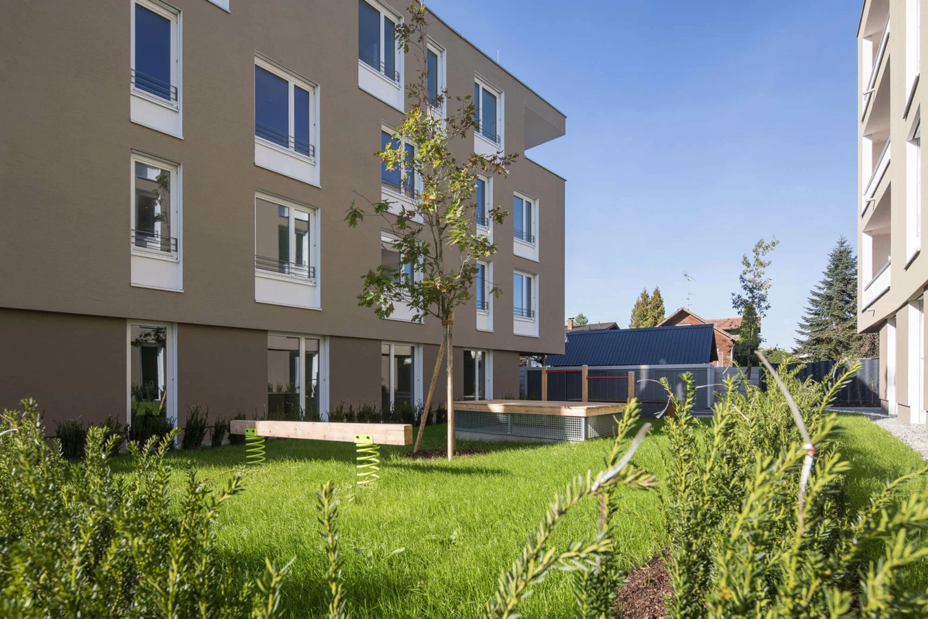 Moderne Wohnanlage Widum Lustenau mit brauner Fassade, vielen Fenstern und einem Spielplatz auf grüner Wiese.