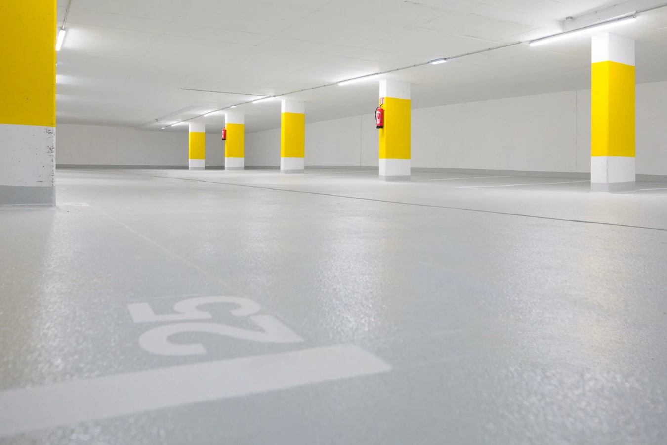 Leere, beleuchtete Tiefgarage mit vielen gelb und weiß gestrichenen Säulen und grauen Stellplätzen, Nummer 25 auf dem Boden (Widum, Wohnanlage, Lustenau).