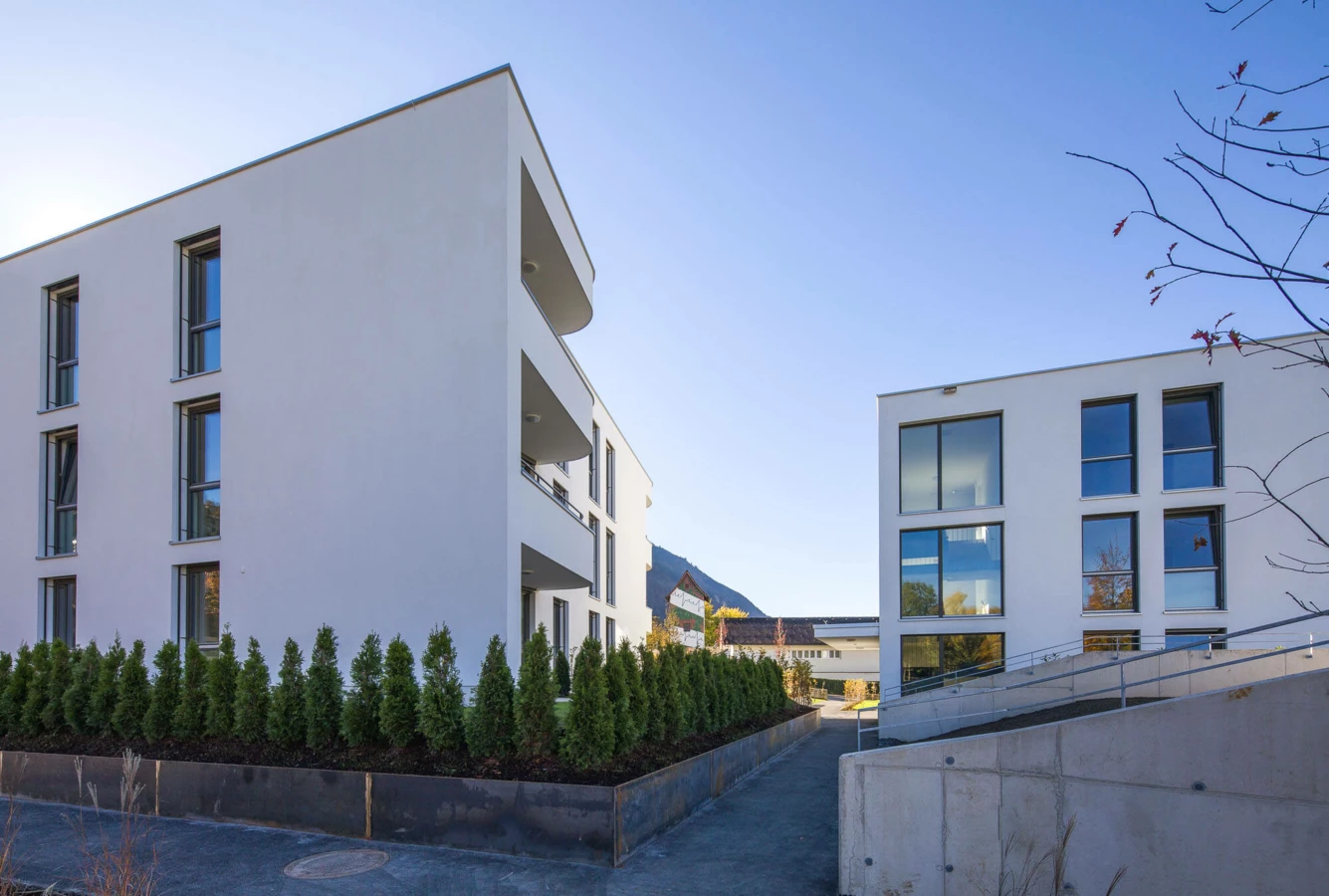 Zwei moderne weiße Wohngebäude mit Balkonen, umgeben von gepflegten Hecken und Gehwegen unter blauem Himmel in der Wolf-Huber-Straße, Feldkirch (Wohnanlage).