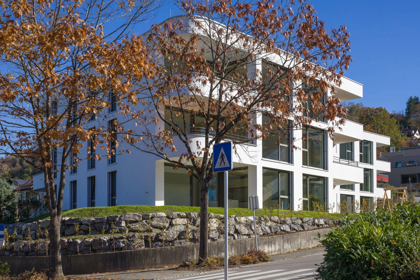 Moderne weiße Wohnanlage an der Wolf-Huber-Straße in Feldkirch mit großen Fenstern und Balkonen, umgeben von Bäumen und einer Steinmauer.