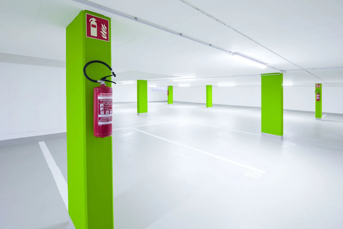 Leere, beleuchtete Tiefgarage mit hellgrünen Säulen, Feuerlöschern und weißen Parkplatzmarkierungen (Wolf-Huber-Straße, Wohnanlage, Feldkirch)