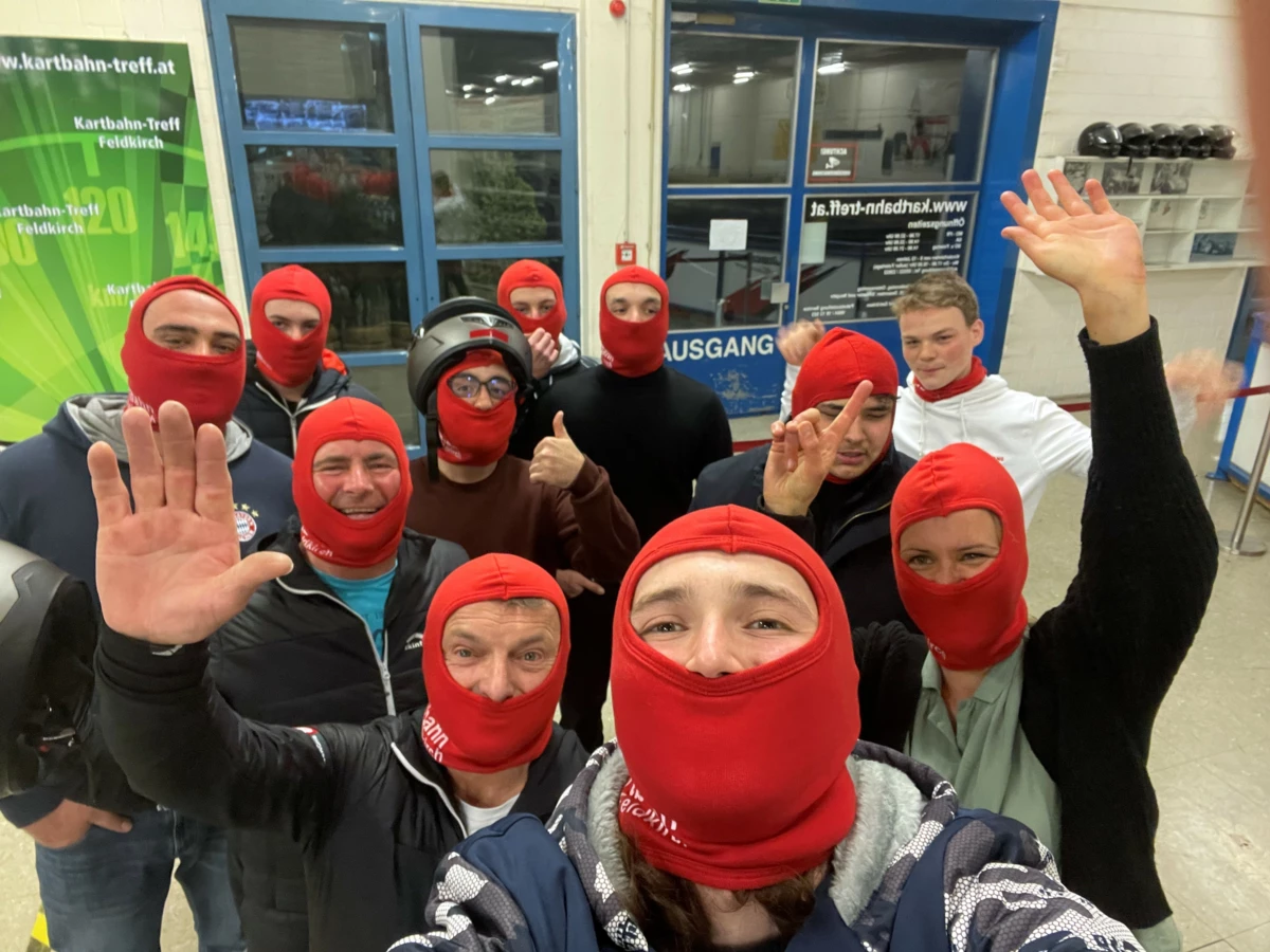 Eine Gruppe fröhlicher Personen trägt rote Sturmhauben und posiert für ein Selfie vor einem Schild mit der Aufschrift "Kartbahn-Treff Feldkirch" und "Ausgang" (Lehrlingsabend).