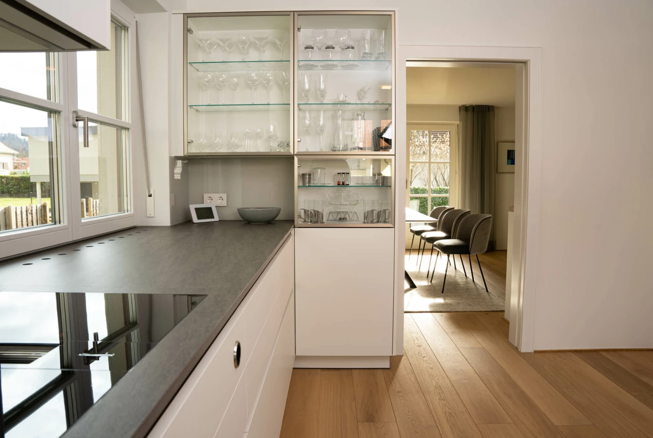 Moderne weiße Küche mit dunkler Arbeitsplatte und Induktionskochfeld, inklusive Glasvitrinenschrank und Blick ins Esszimmer mit Holzboden (Generalsanierung, Bohle).