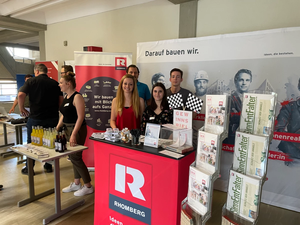Messestand des Bauforums mit Rhomberg Bau Beschriftung, Informationsmaterialien und Mitarbeitern, die bereitstehen, die Besucher des Messestands zu begrüßen.