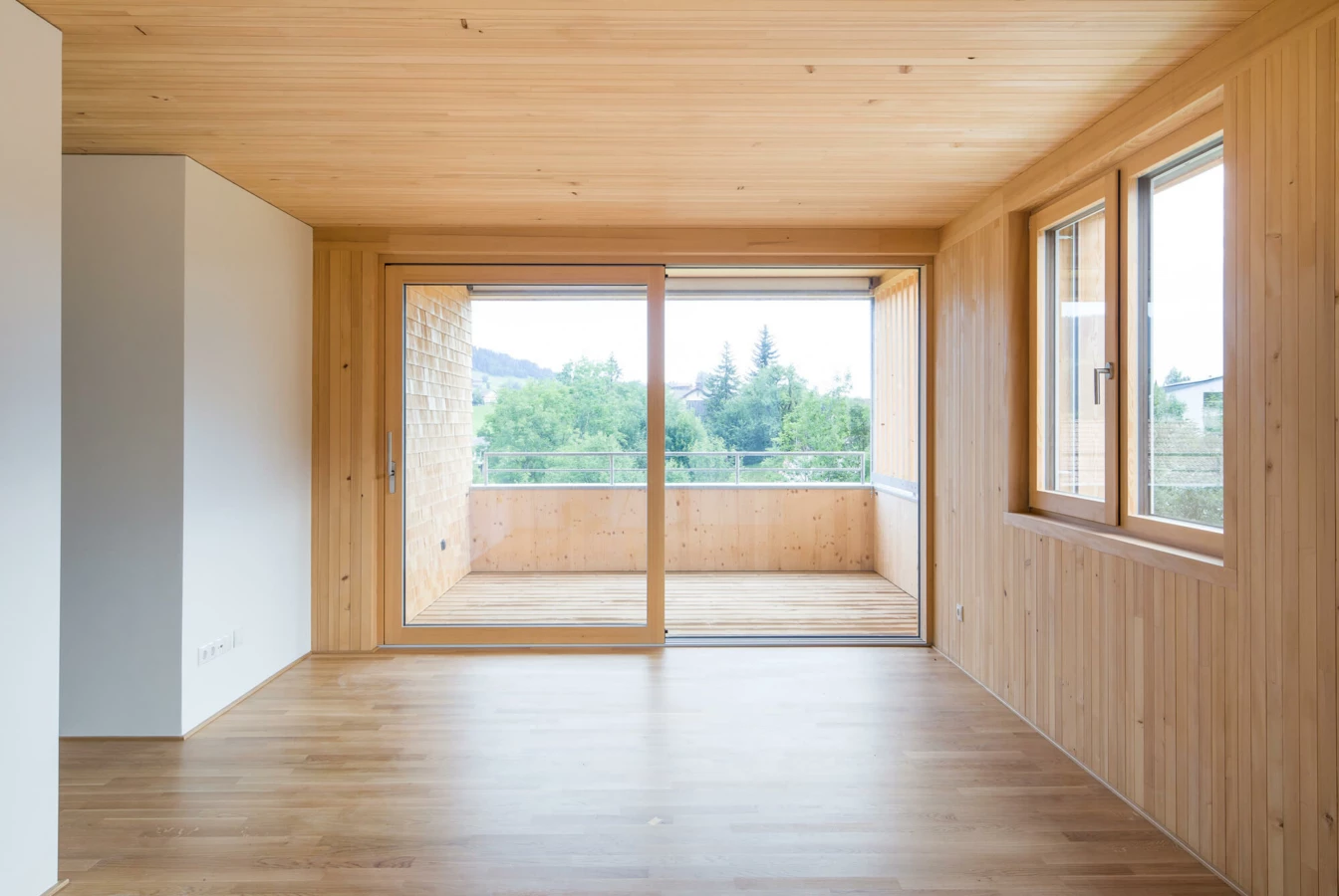 Leerer Raum mit Holzboden und Holzdecke, grosser Schiebetür zum Balkon und Fenster mit Blick ins Grüne (Wohnanlage, Alberschwende)