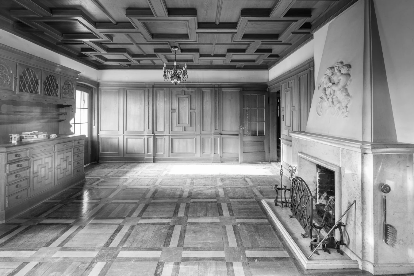 Schwarz-Weiß-Ansicht eines leeren Raumes mit Holzpaneelen, Kassettendecke, Kamin und Parkettboden, dazu ein Kronleuchter (Villa, Reichsstr).