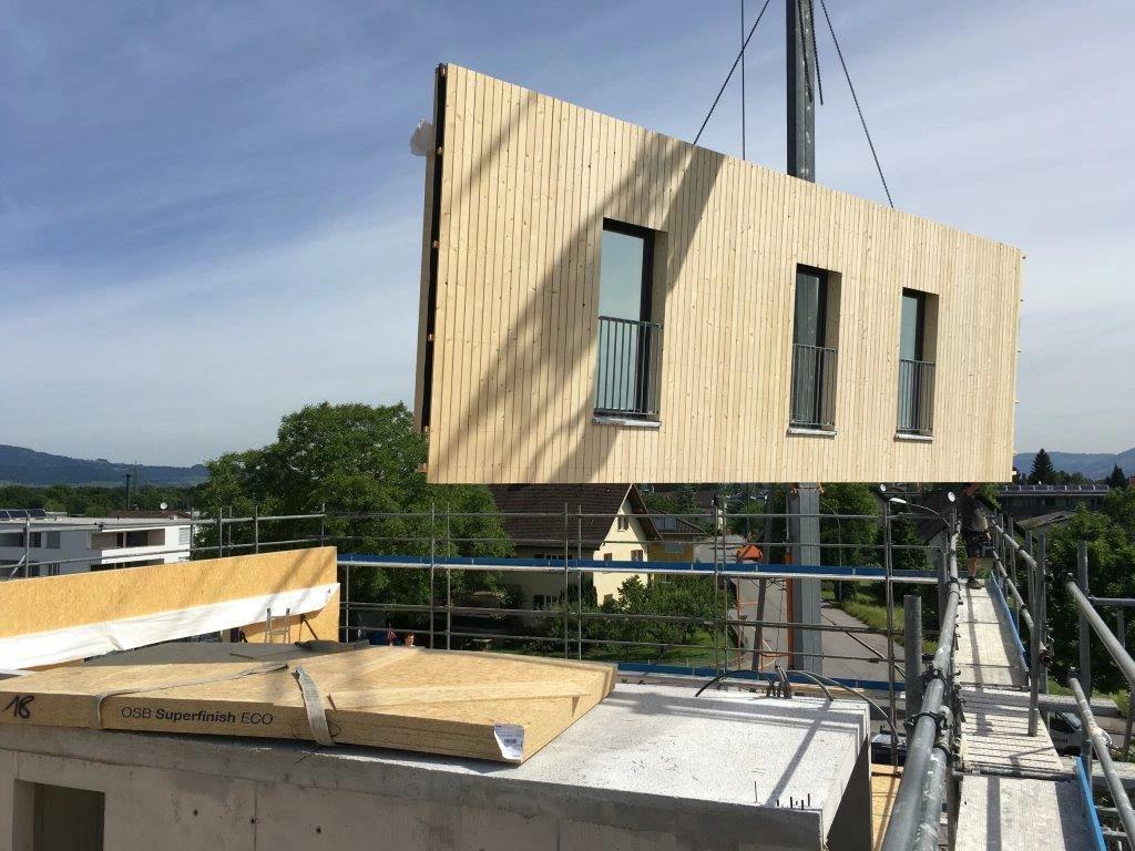 Ein Kran hebt ein vorgefertigtes Holzbaumodul mit drei Fenstern und Balkonen auf eine Baustelle (Architekturexkursionen, Holzbauprojekte)