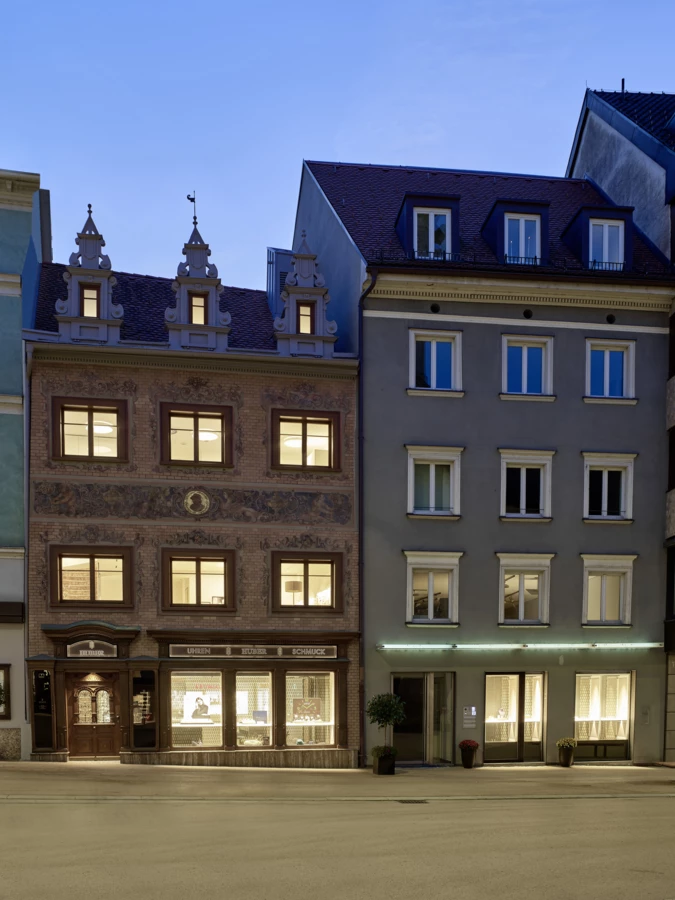 Zwei beleuchtete mehrstöckige Gebäude in der Rathausstraße 7 während der Dämmerung, eines mit 'Uhren Huber' Beschriftung und Schaufenstern.