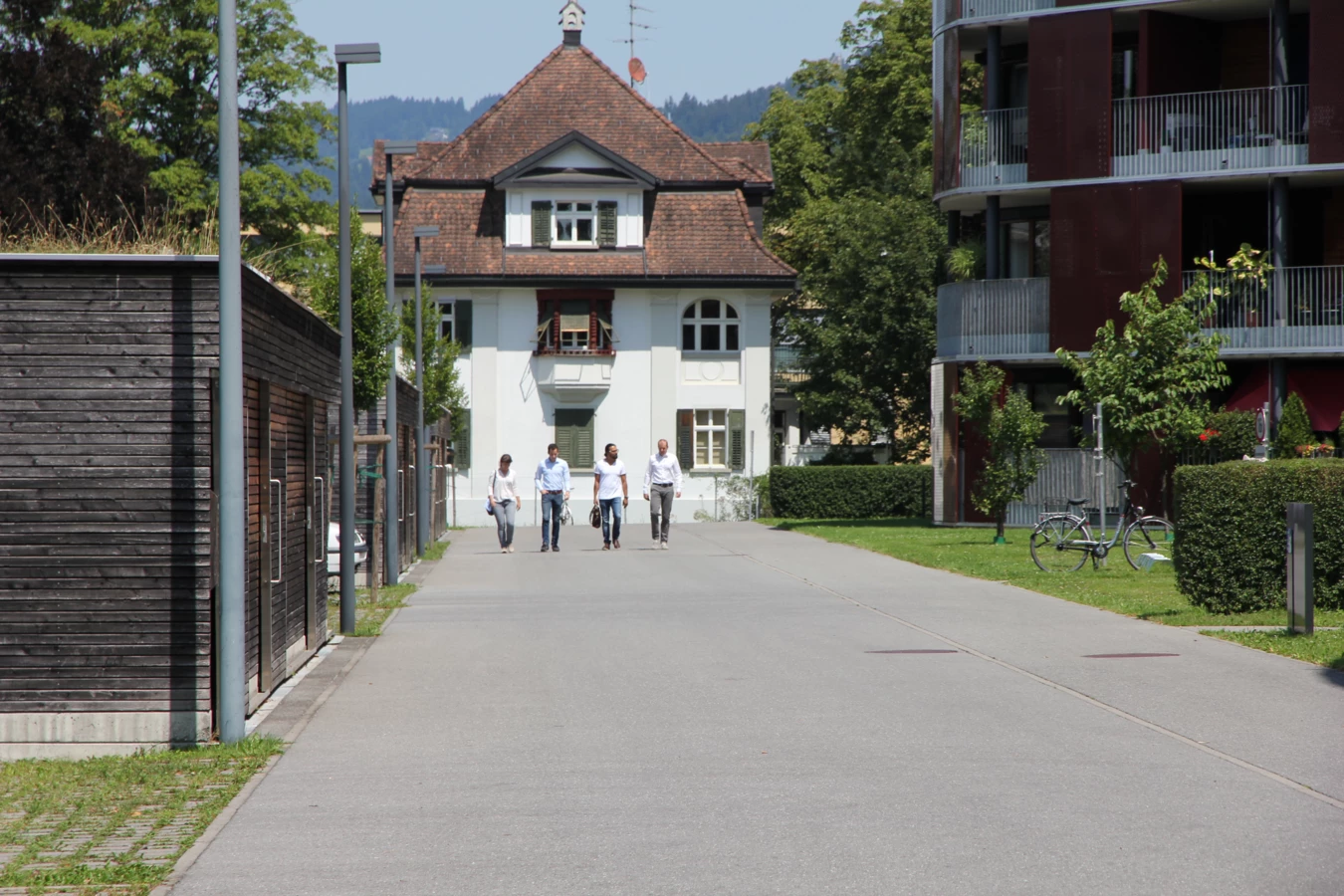 Vier Personen gehen eine Straße entlang, mit einem traditionellen weißen Haus und modernen Gebäuden im Hintergrund.