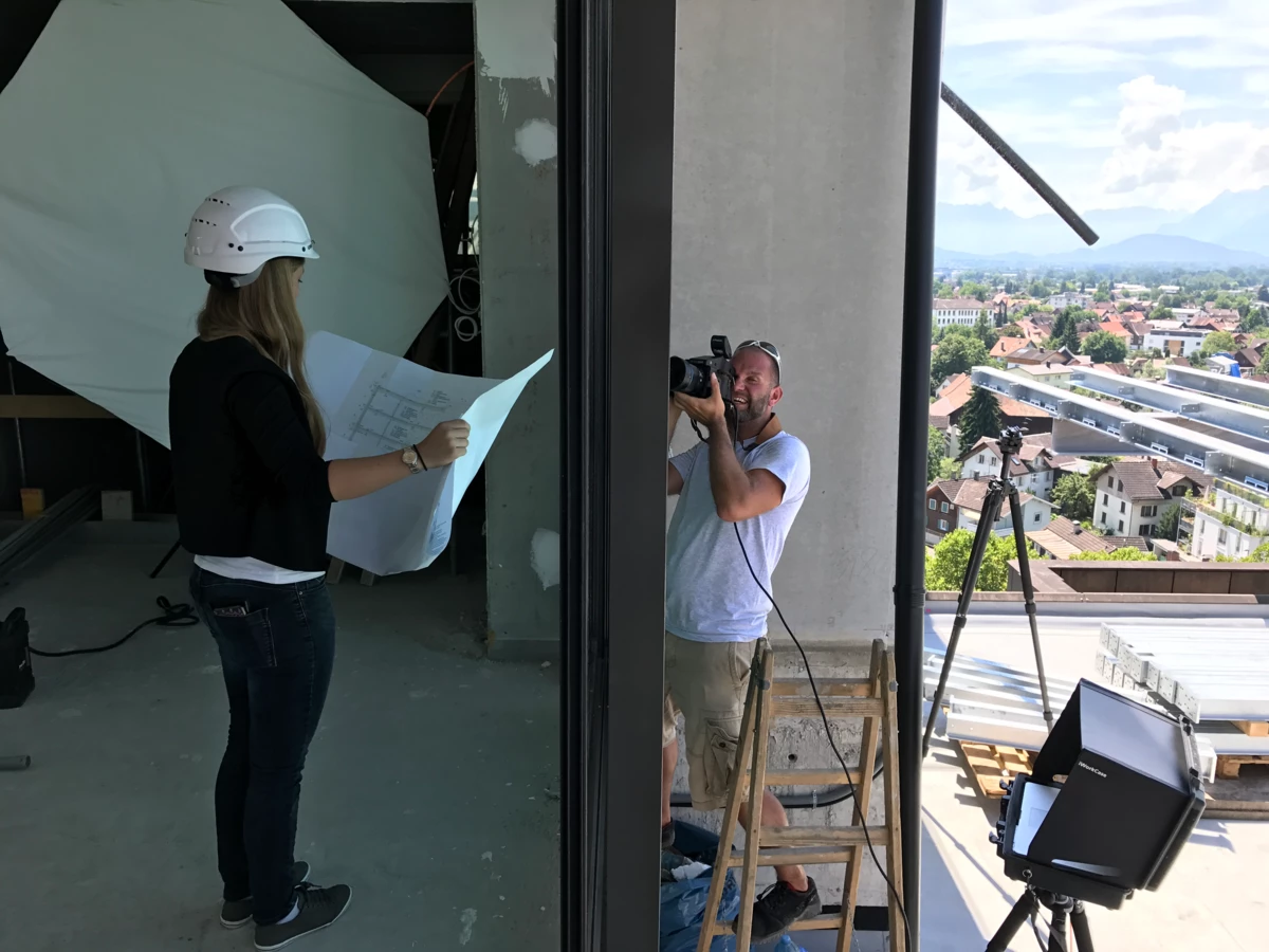 Eine Frau mit Schutzhelm und Bauplänen sowie ein Fotograf auf einer Leiter mit Kamera bei einem Fotoshooting auf einer Baustelle.