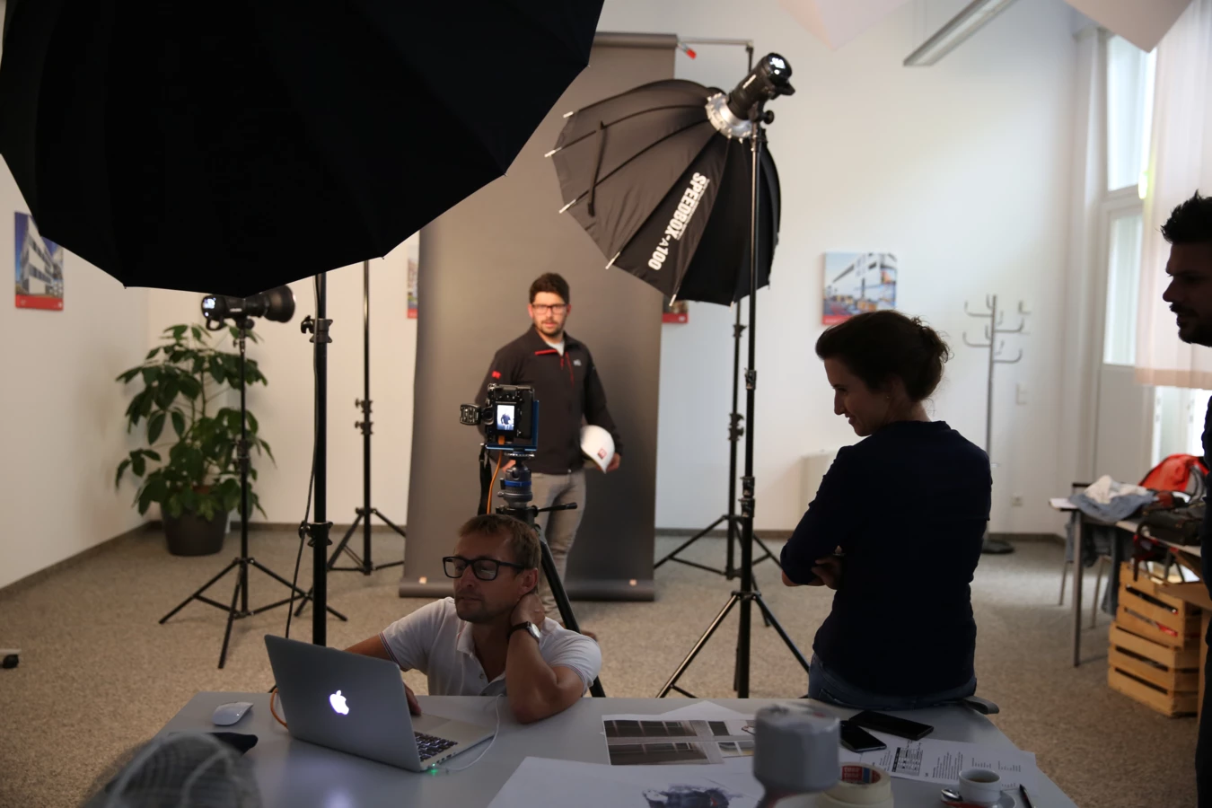 Männer und Frauen bei einem Fotoshooting, ein Mann sitzt vor einem Laptop, während ein anderer Mann posiert und eine Frau zusieht.