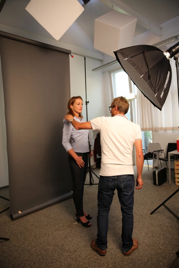 Ein Fotograf positioniert eine Frau vor einem grauen Hintergrund während eines Fotoshootings im Innenbereich.