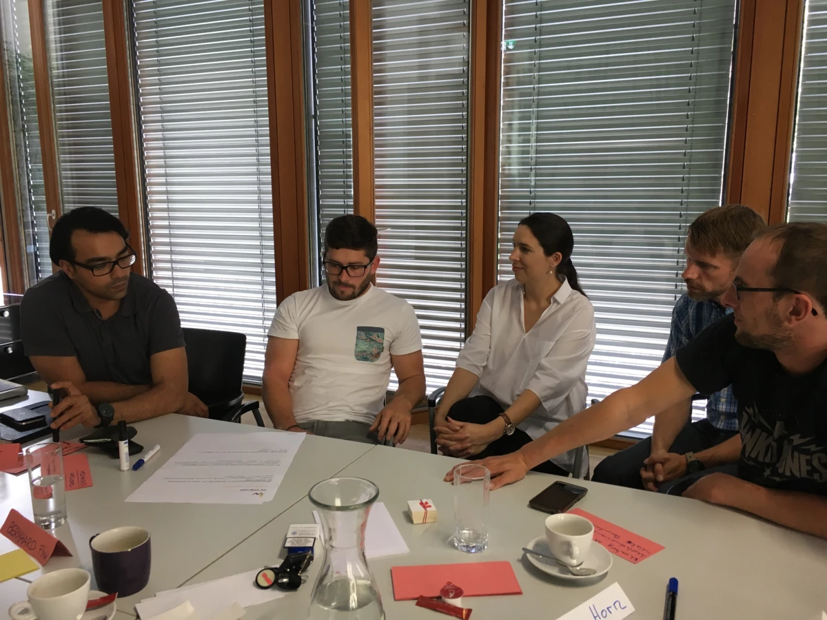 Fünf Personen sitzen an einem Tisch, besprechen Dokumente und trinken Kaffee in einem Workshop-Umfeld bei Rhomberg Bau.