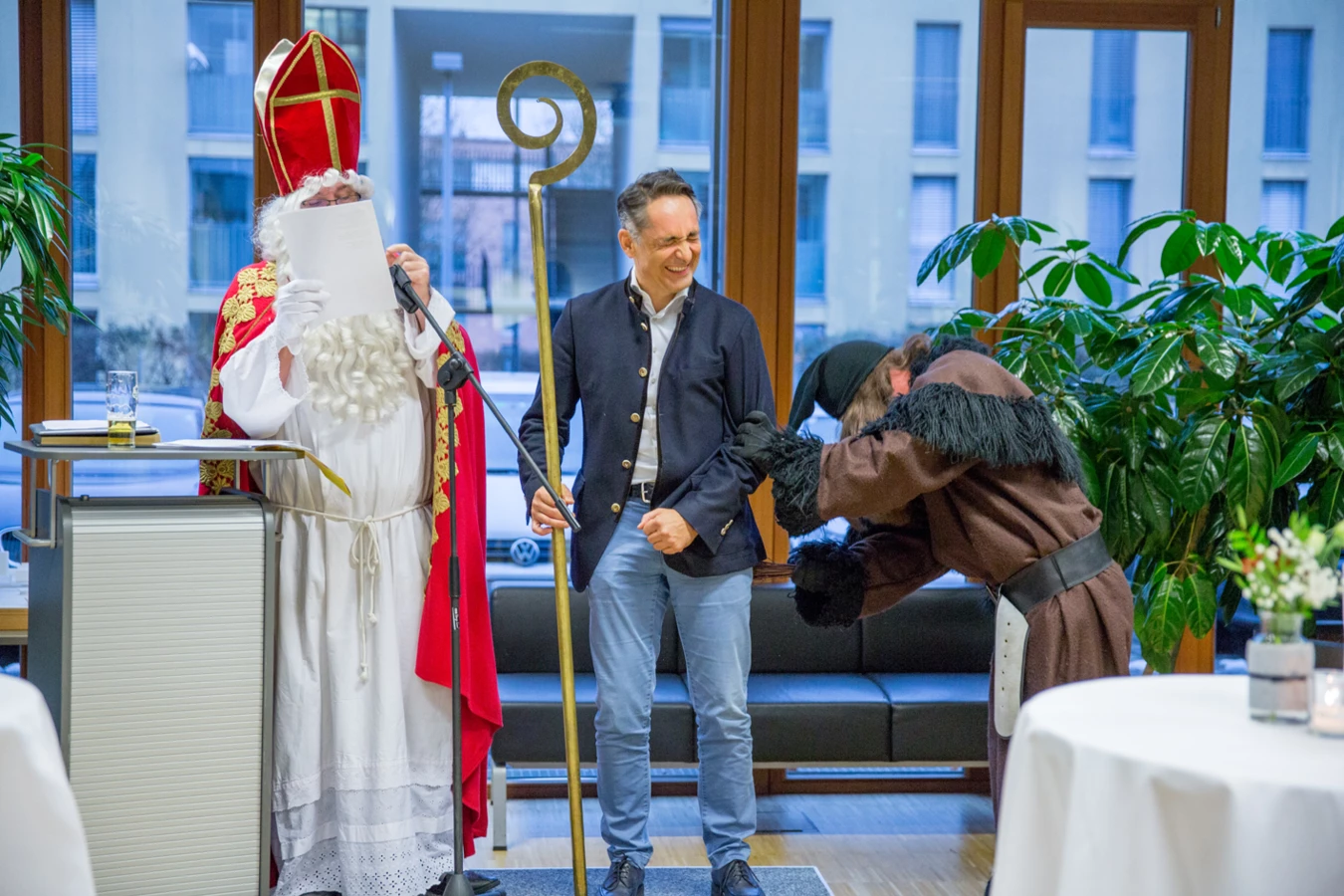 Ein Mann im Nikolaus-Kostüm liest von einem Blatt ab, während ein anderer Mann lachend einen Bischofsstab hält und ein Krampus zu seinen Füßen kauert (Hubert, Geburtstag).