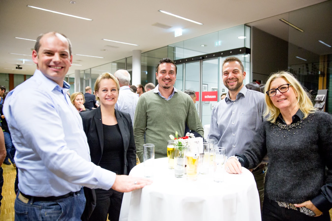 Fünf lächelnde Personen stehen um einen runden Tisch mit Getränken und Blumen bei einer Veranstaltung (Hubert, Geburtstag)