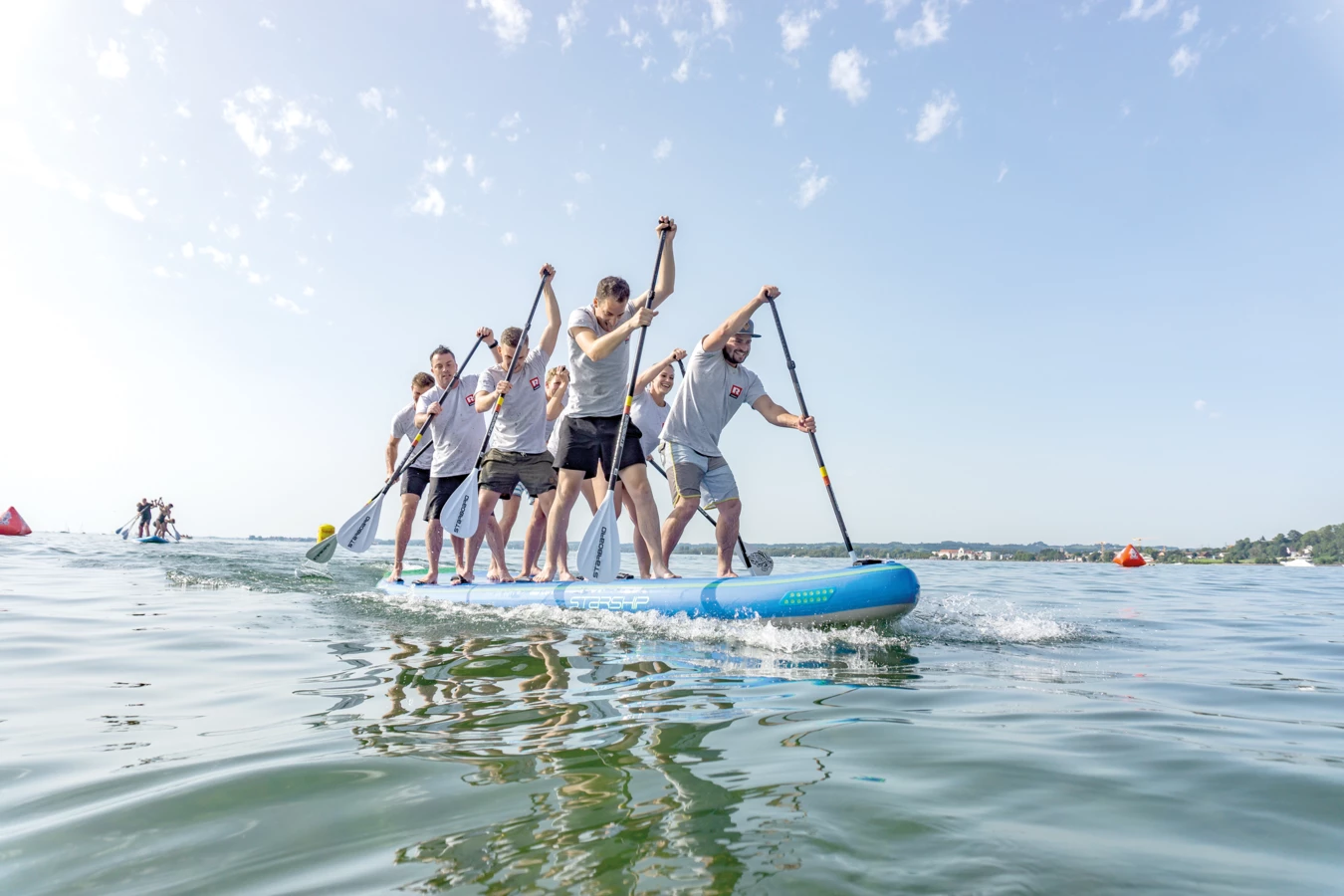 Eine Gruppe von sechs Personen paddelt gemeinsam stehend auf einem großen blauen SUP-Board über klares Wasser unter strahlend blauem Himmel (MathiasJoschika).