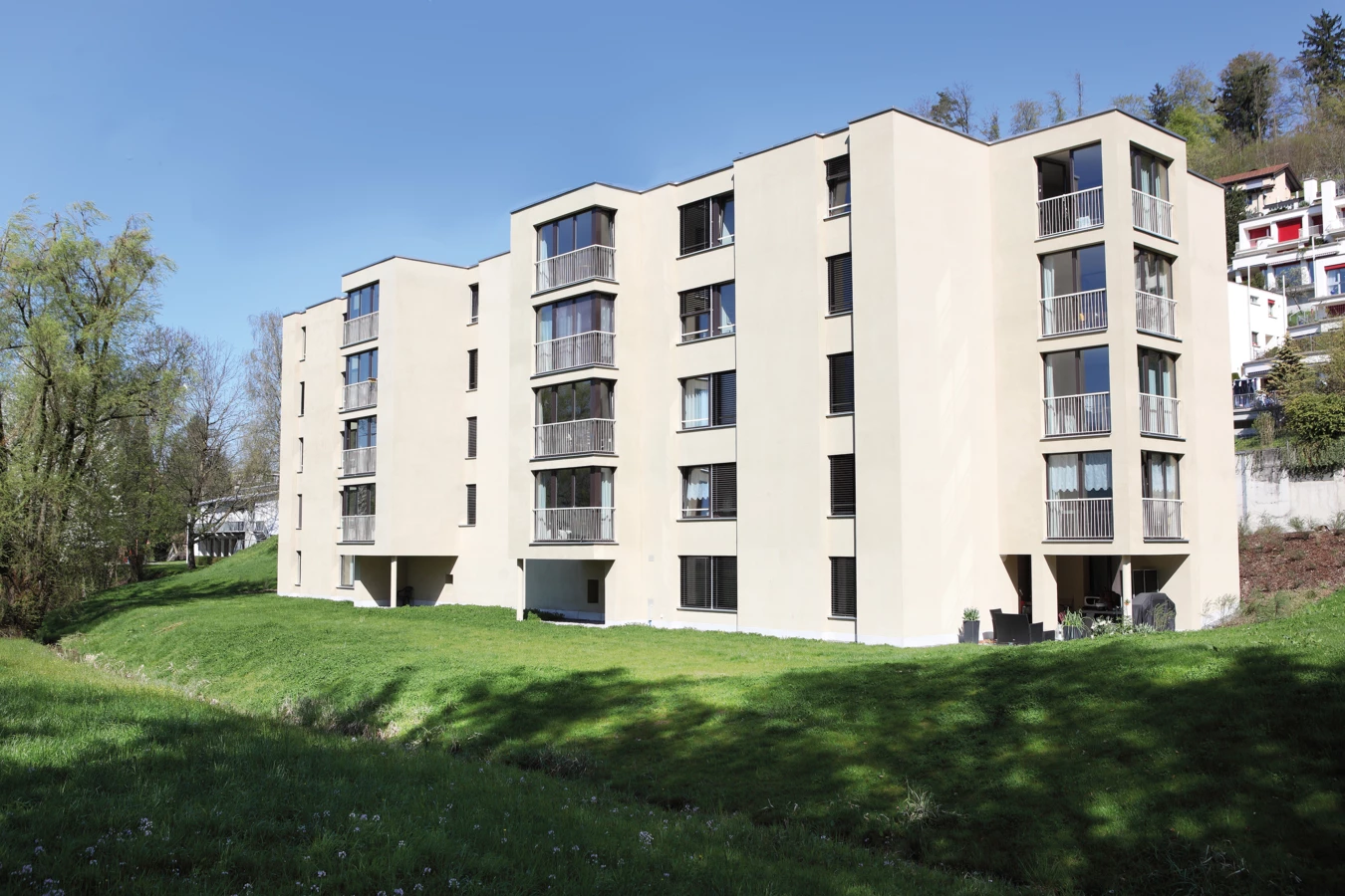 Modernes beige Mehrfamilienhaus mit Balkonen und Jalousien, umgeben von grünen Wiesen und Bäumen unter blauem Himmel