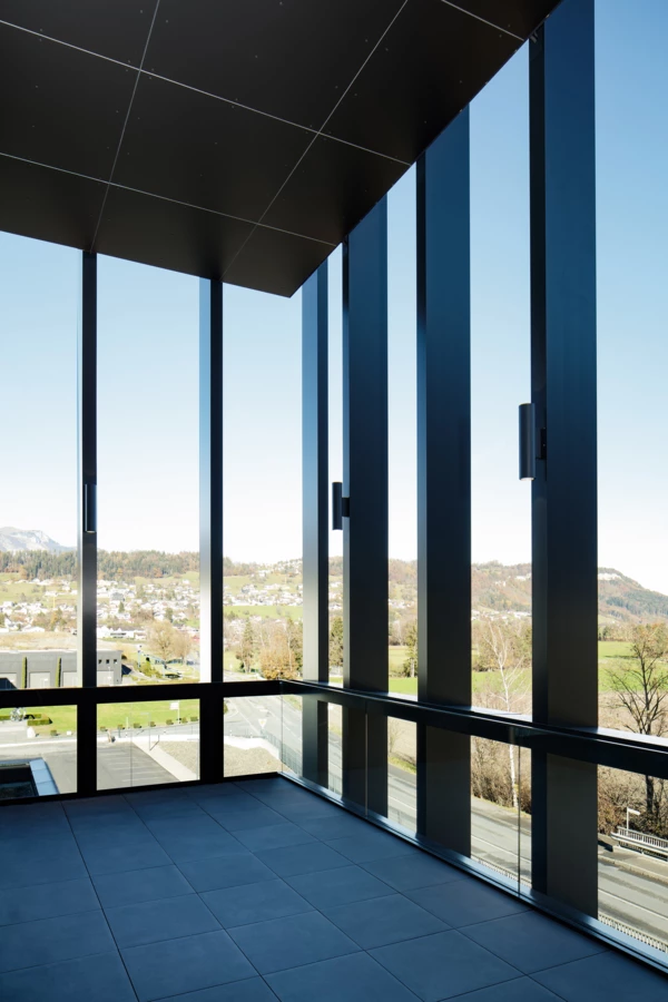 Ein moderner Raum mit großen Fenstern, die den Blick auf ein grünes Tal mit Häusern und Bergen freigeben. Schwarze Fliesen auf dem Boden (Edenta).