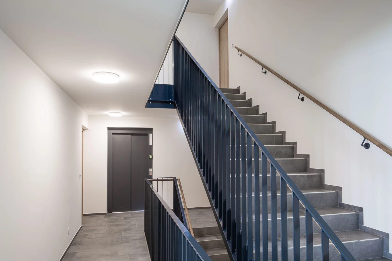 Modernes Treppenhaus mit grauem Fliesenboden, dunklem Metallgeländer, hellem Holzhandlauf und einem Aufzug in der Wohnanlage Bahnhofsstrasse Vandans (Stiegenhaus).