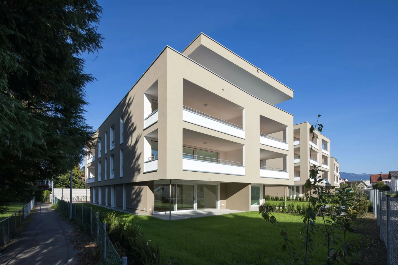 Moderne Wohnanlage Widum Lustenau mit beigefarbenen Fassaden und Balkonen, umgeben von Grünflächen und einem Fußweg unter klarem Himmel.